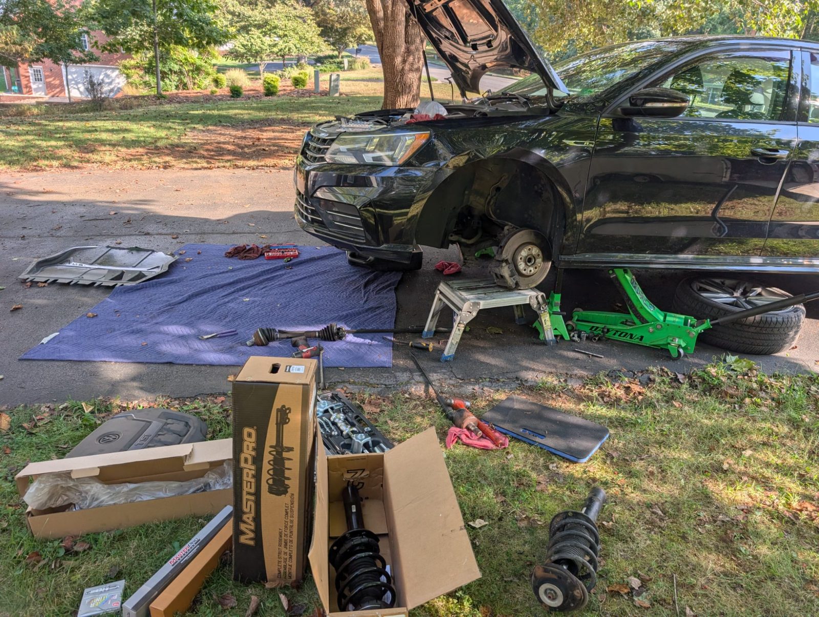 Black car on a jack in a driveway with new struts, axle and tools spread on a mat