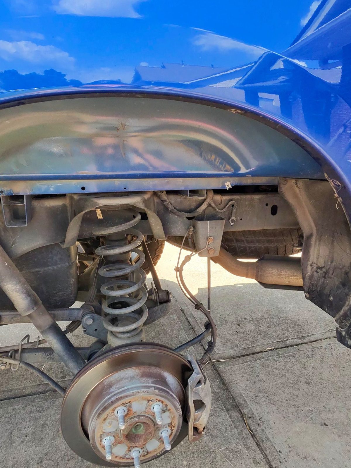 Front wheel removed from a blue vehicle in a driveway, showing brake rotor, hub and coil spring