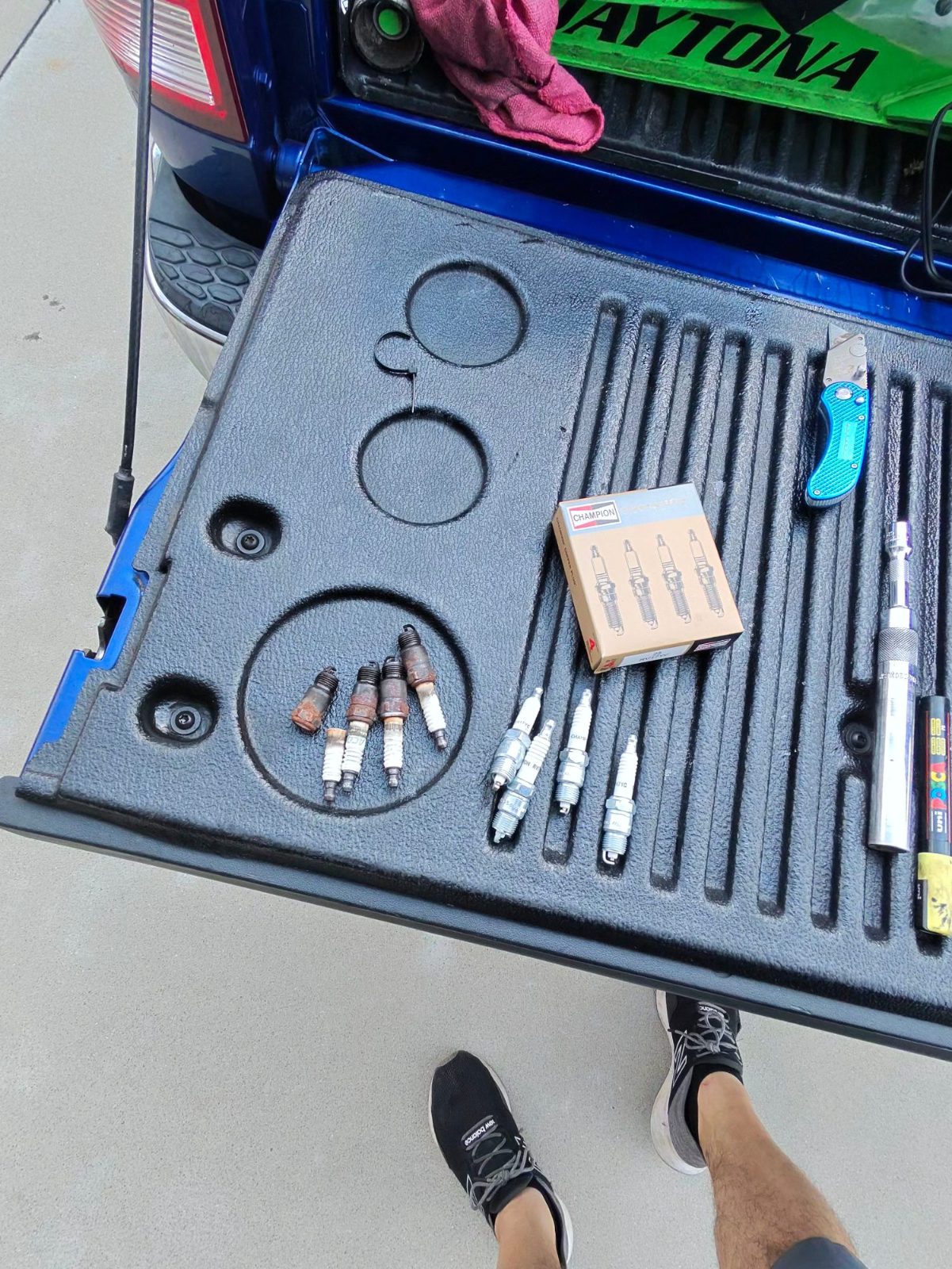 Old and new spark plugs laid out on a truck tailgate beside a box and hand tools