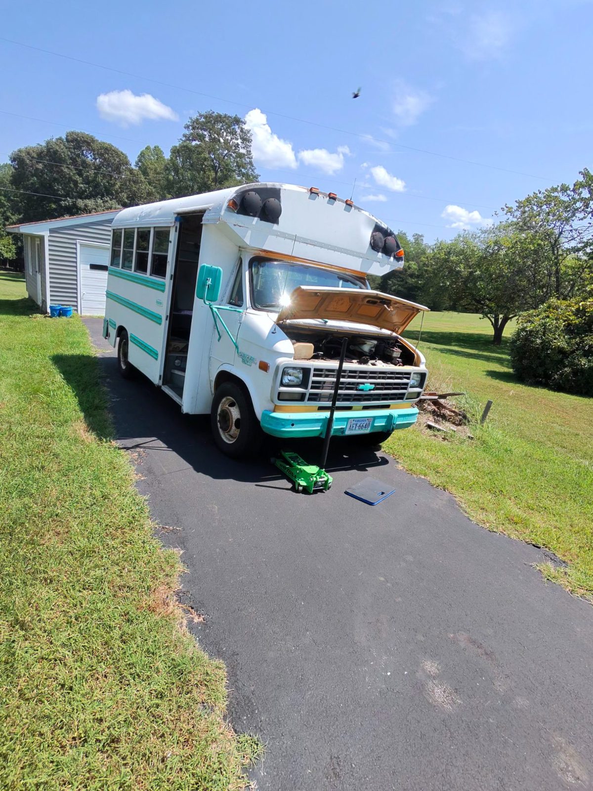 Small white and teal bus with its hood up in a driveway, floor jack under the front bumper